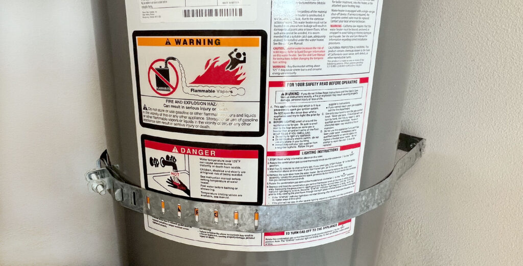 Water heater with safety warning labels and a metal strap for earthquake protection.